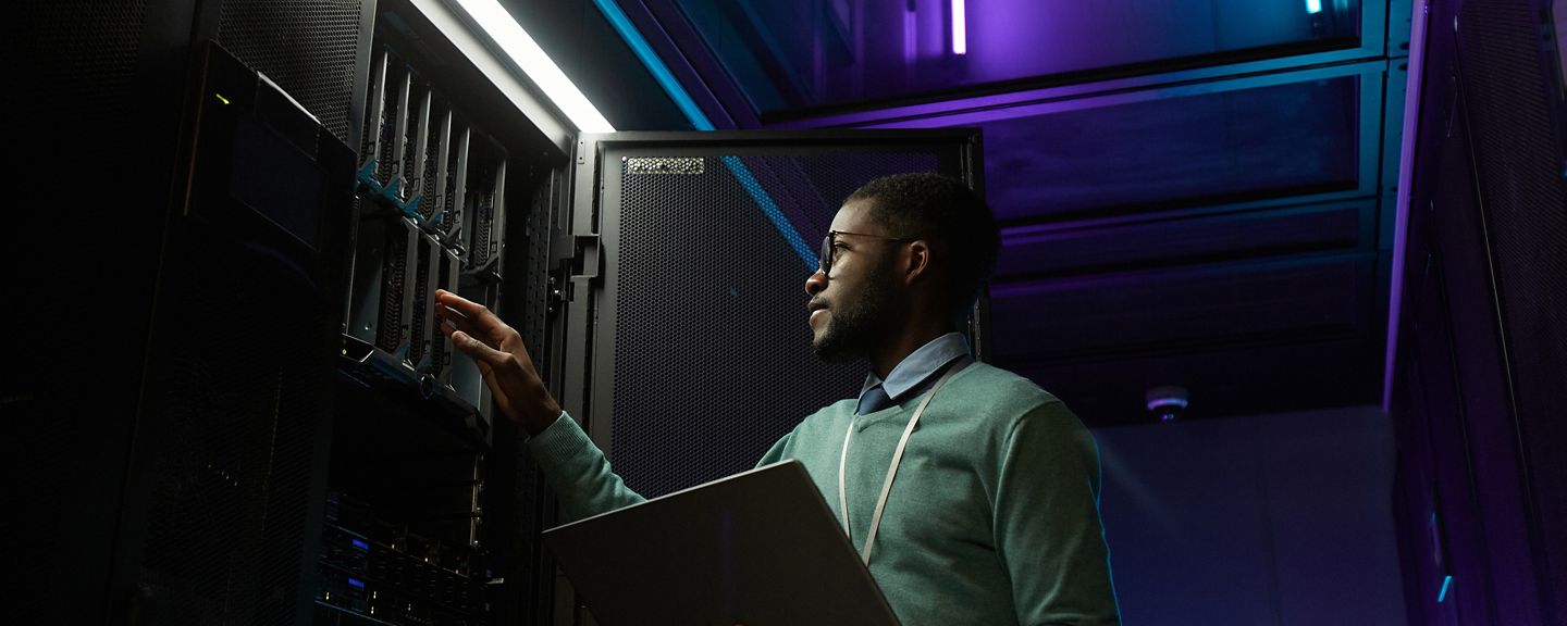 Low angle portrait of young African American data engineer working with supercomputer in server room lit by blue light and holding laptop, copy space