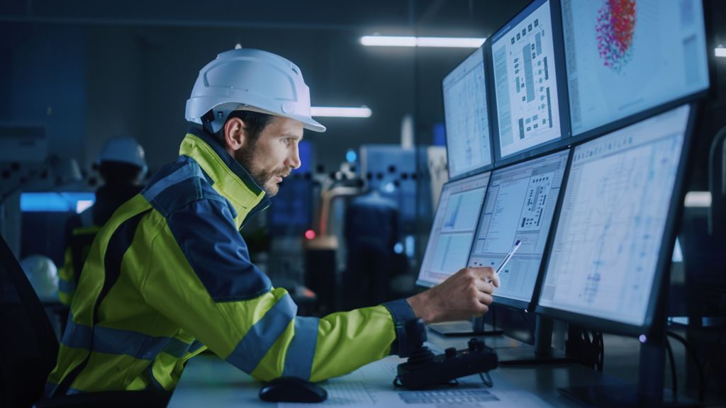 Industry 4.0 Modern Factory: Facility Operator Controls Workshop Production Line, Uses Computer with Screens Showing Complex UI of Machine Operation Processes, Controllers, Machinery Blueprints