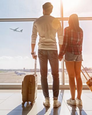 Inspired young loving couple is looking at flying plain in sky. They are standing near window at airport and holding hands