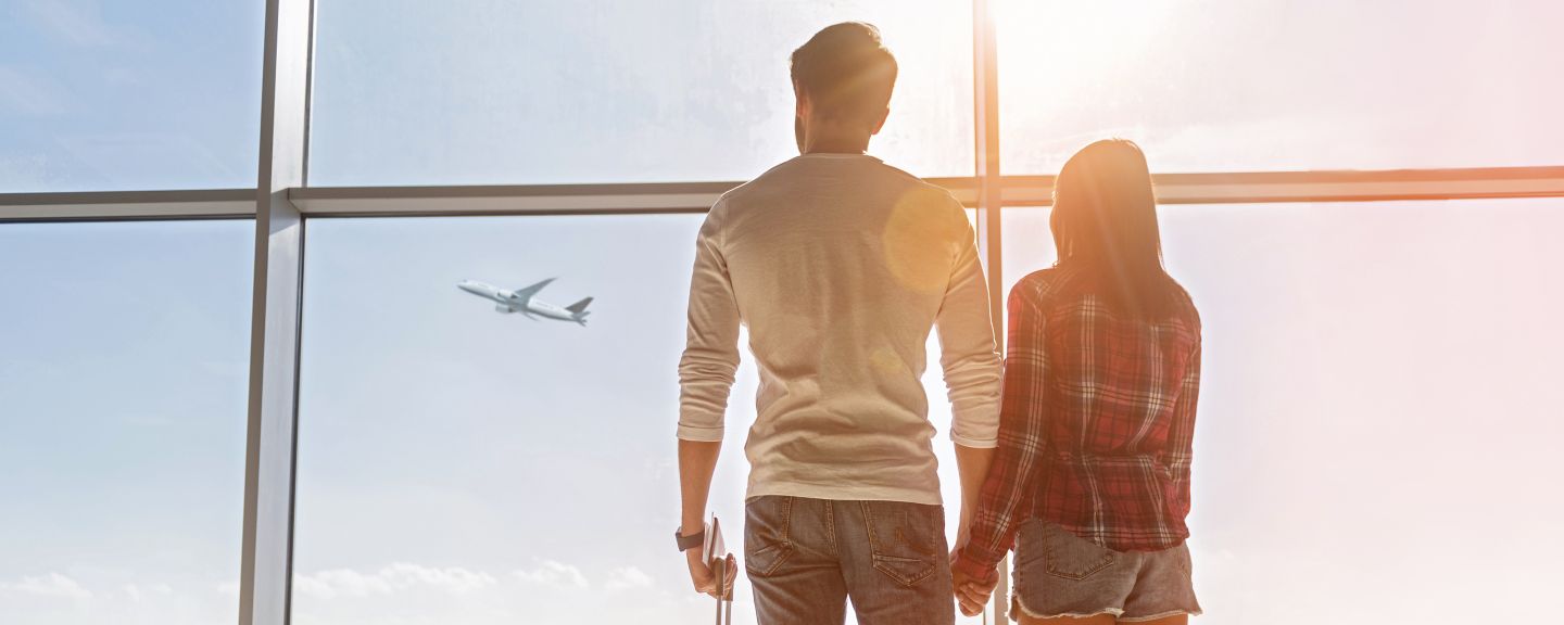 Inspired young loving couple is looking at flying plain in sky. They are standing near window at airport and holding hands