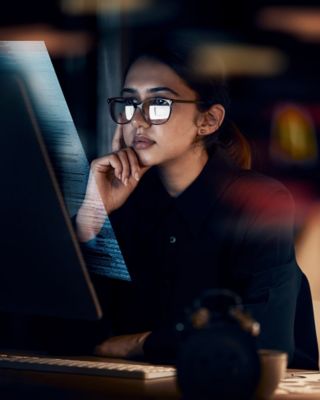 Focused professional woman wearing glasses, analyzing information on a computer screen in a dimly lit setting.