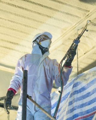 Worker in protective gear spraying insulation foam on a ceiling inside a construction site, representing energy-efficient building insulation.