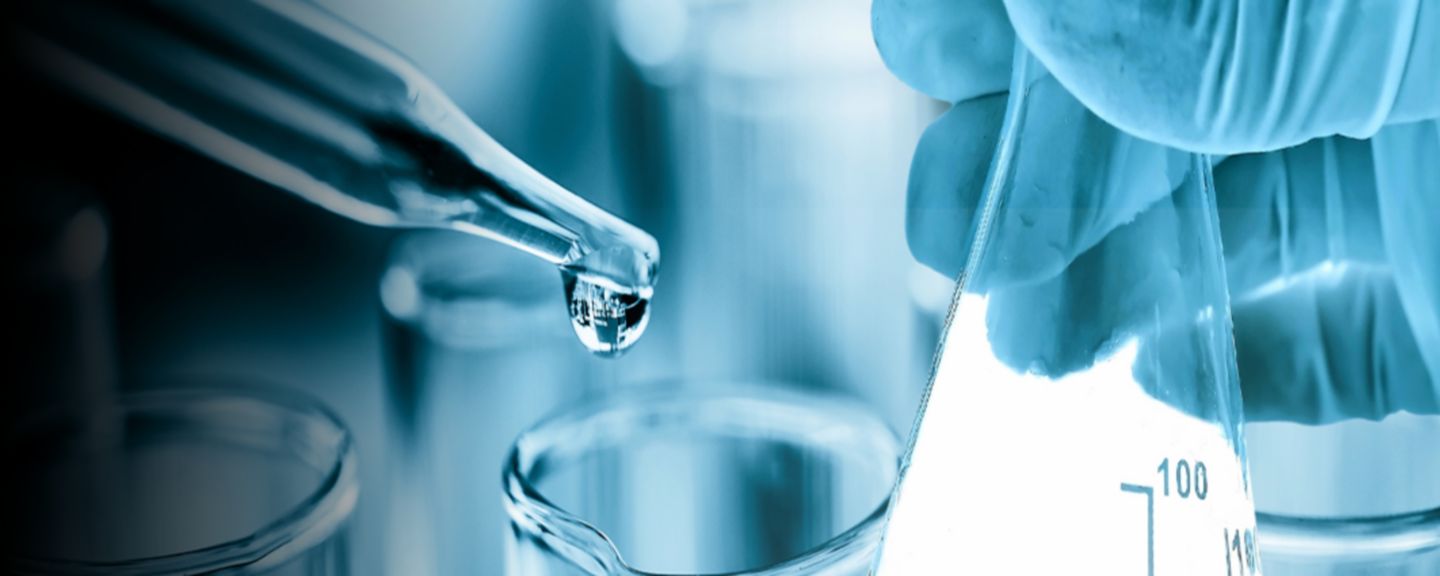 Close-up of a scientist’s gloved hands holding a laboratory flask as a pipette releases a drop of liquid, surrounded by glass test tubes in a scientific research lab.