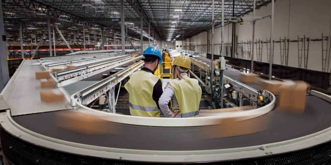 Two supervisors in safety gear observe a busy warehouse with conveyor belts and packages in motion.
