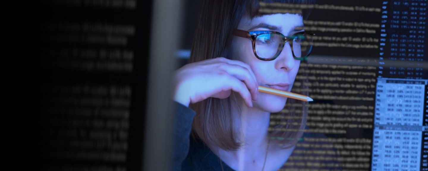 Focused professional woman wearing glasses working on a computer surrounded by lines of code, representing digital talent and career opportunities in technology.
