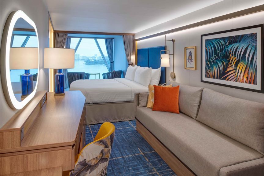 Modern room with a king bed, gray sofa, blue accents, colorful artwork, and a waterfront city view through large windows.