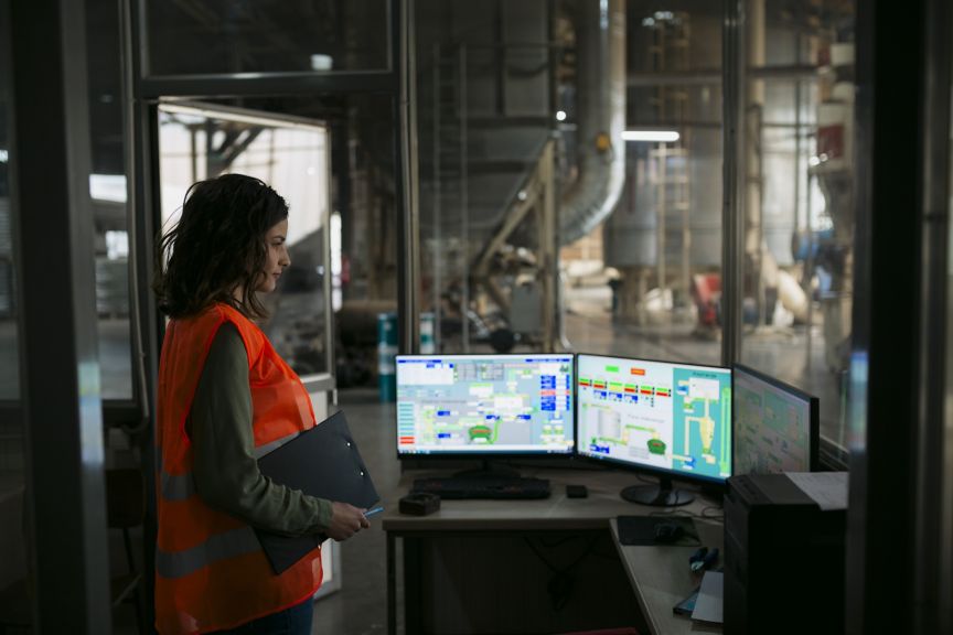 Female industrial worker operating machine on plant floor, looking at monitors over control panel. Worker looking at computer monitors showing automatic production system in wood factory.