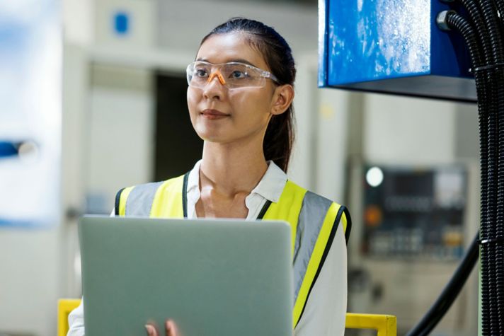 A worker with a safety vest holds a laptop in an industrial plant or manufacturing environment.