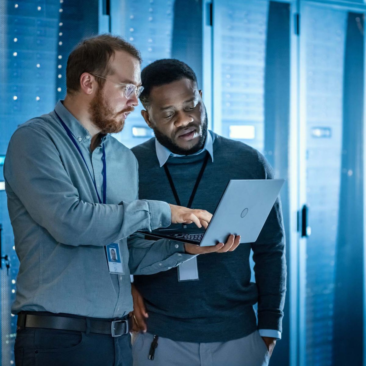 Bearded IT Technician in Glasses with Laptop Computer and Black Male Engineer Colleague are Using Laptop in Data Center while Working Next to Server Racks. Running Diagnostics or Doing Maintenance Work