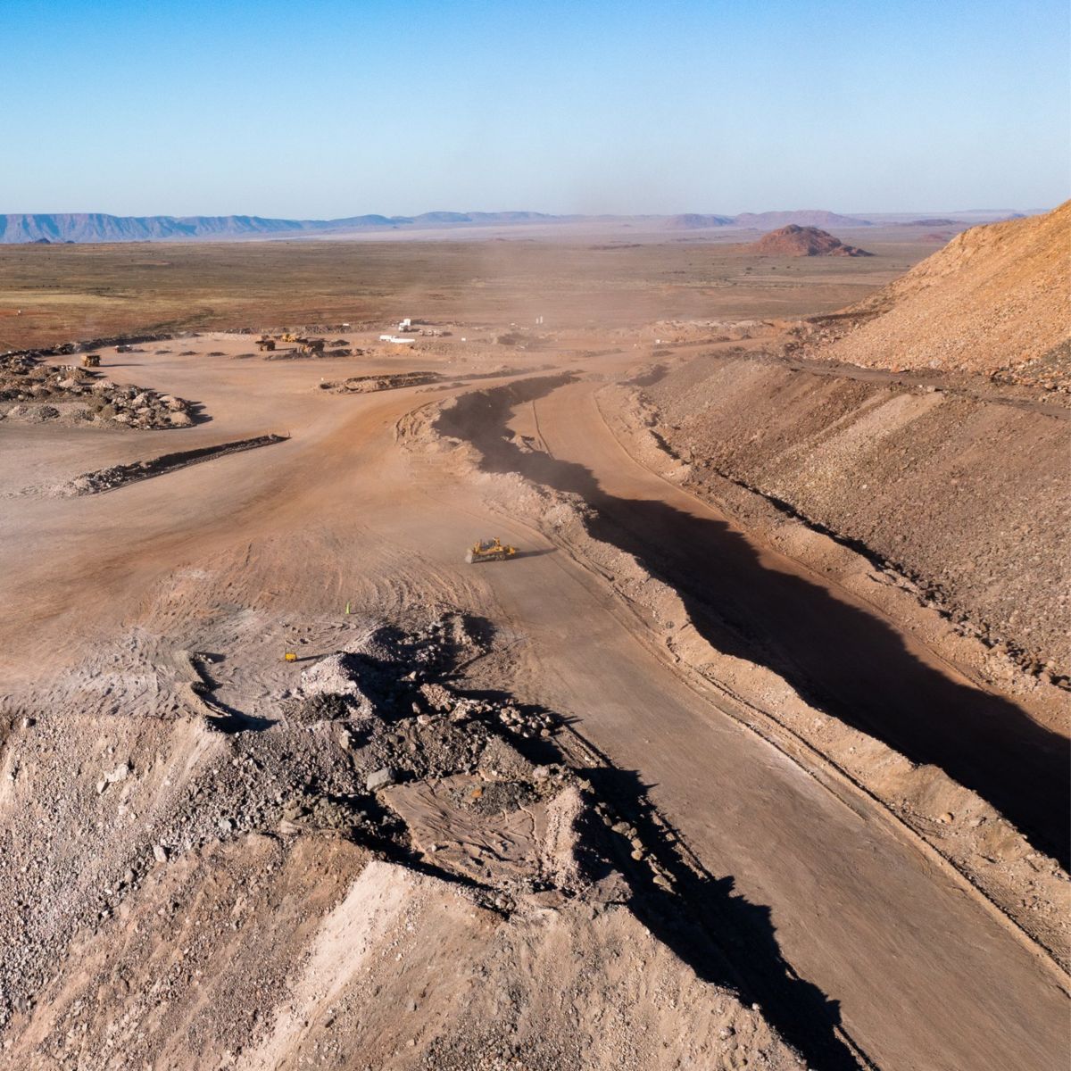 Heavy machinery operates at an open-pit mine site against a vast desert landscape under a clear sky.