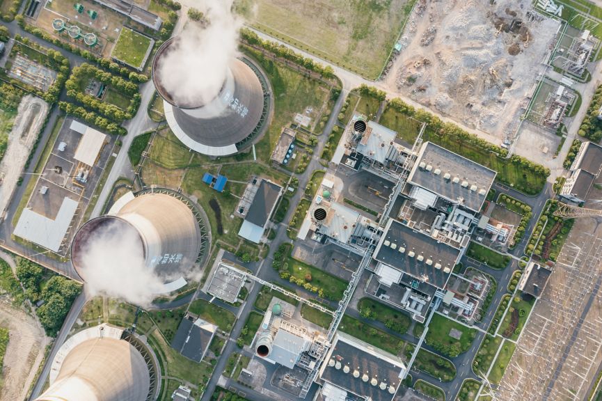 An overhead aerial view shows cooling towers emitting steam alongside a complex industrial power plant facility set amidst green spaces and surrounding land.
