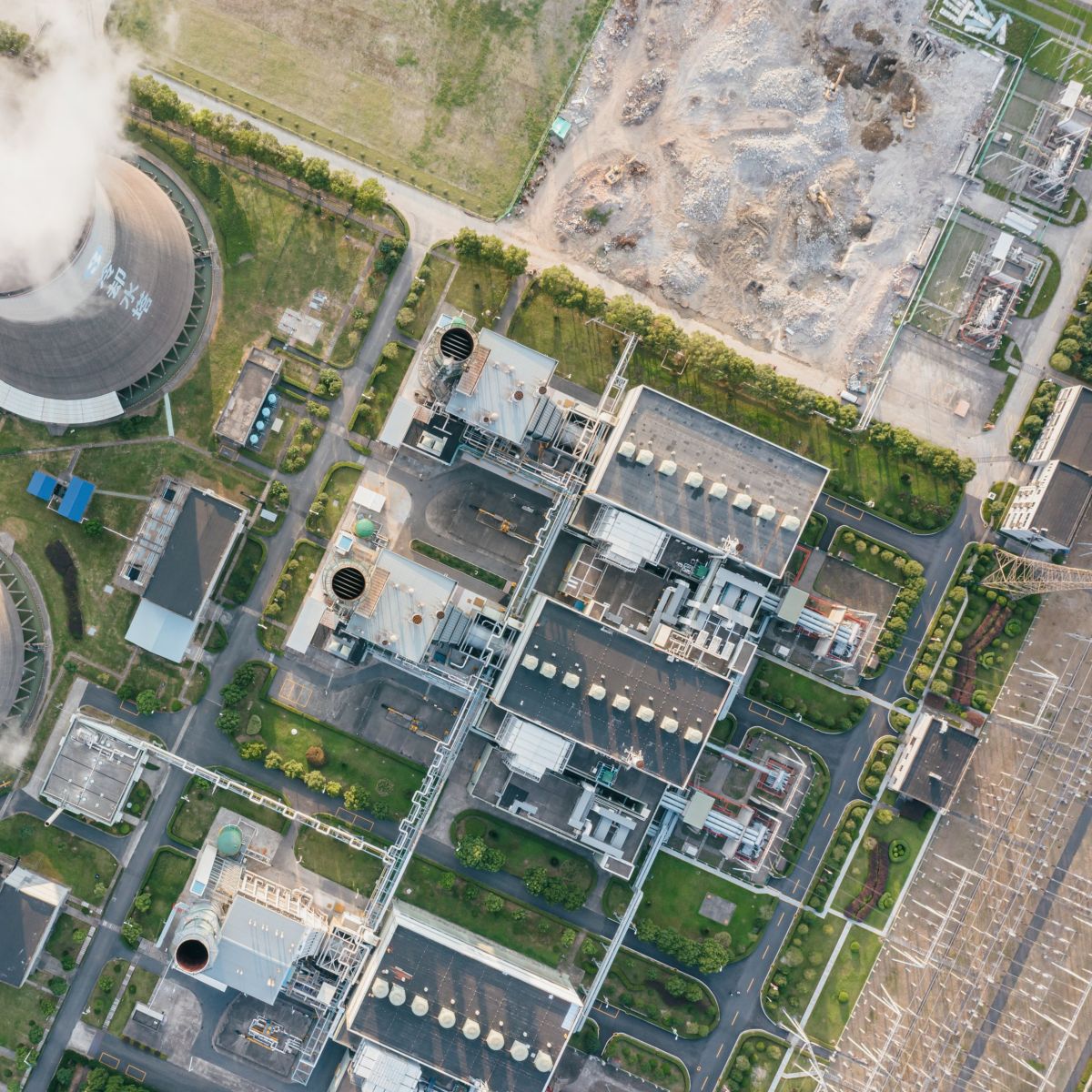 An overhead aerial view shows cooling towers emitting steam alongside a complex industrial power plant facility set amidst green spaces and surrounding land.