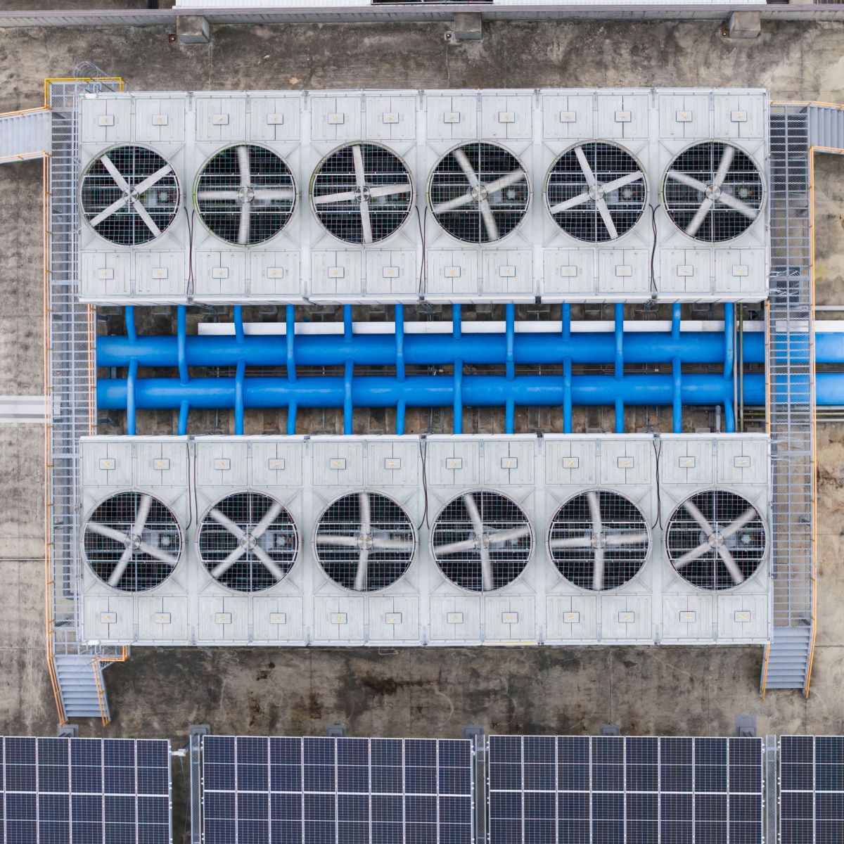 Top view of rooftop cooling units with multiple circular fans, blue piping, and solar panels on a concrete surface.