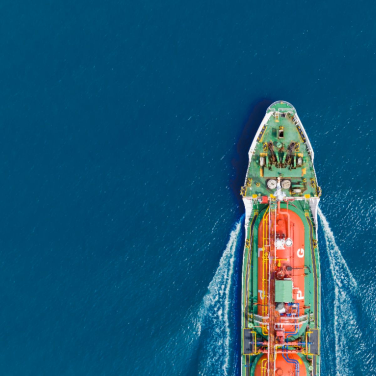 Top‑down view of a green and red tanker ship creating white waves as it moves through blue ocean water.