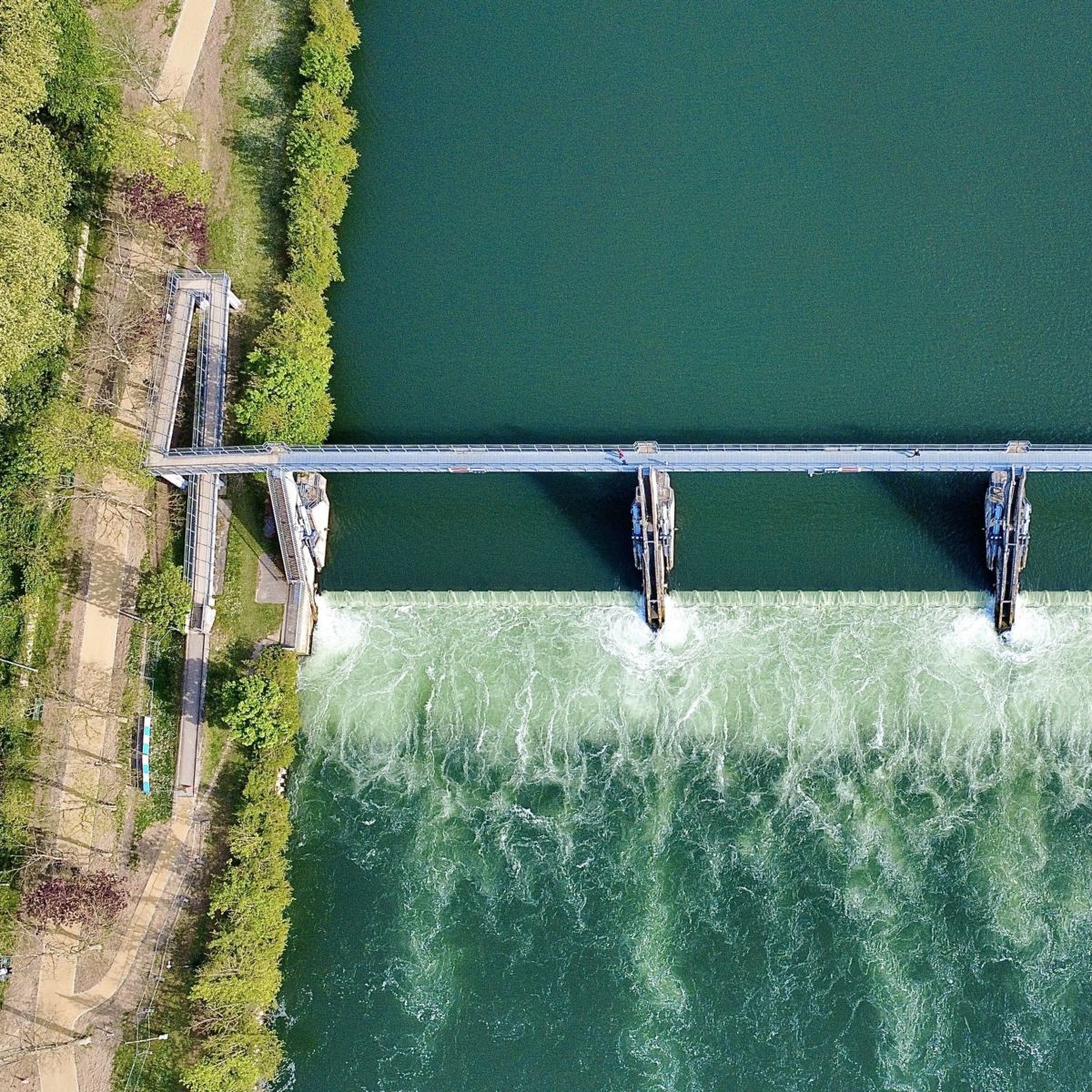 An overhead aerial view shows water flowing over a concrete weir structure on a canal, framed by green riverbanks and a pedestrian access bridge.