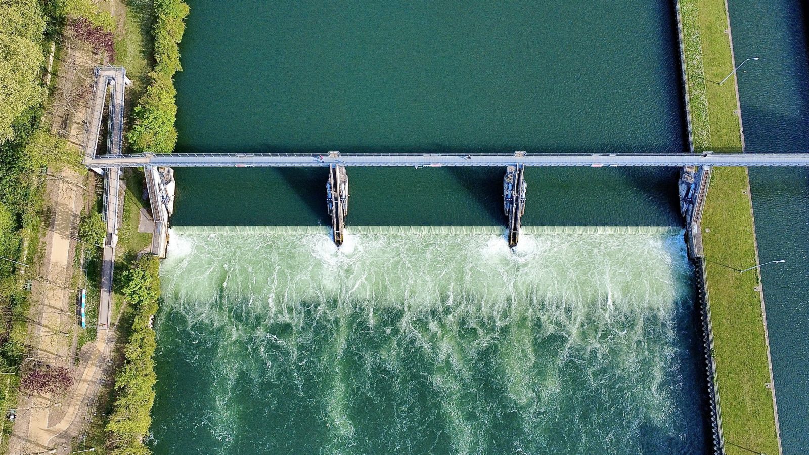 An overhead aerial view shows water flowing over a concrete weir structure on a canal, framed by green riverbanks and a pedestrian access bridge.