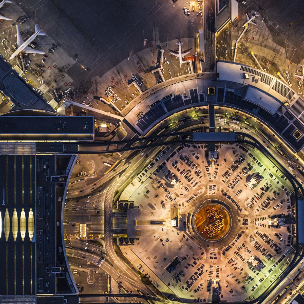 An illuminated aerial view capturing the intricate night-time layout of airport terminals, aircraft parking aprons, and multi-level traffic infrastructure.
