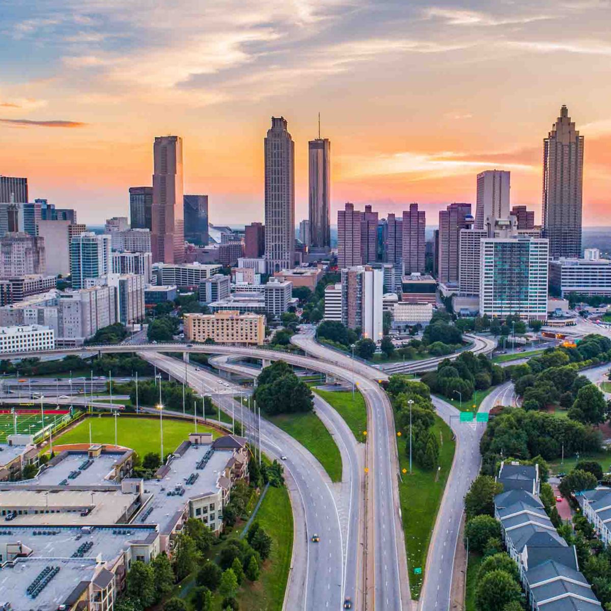 Aerial view of Atlanta’s skyline at sunset, featuring modern skyscrapers and highways amid lush greenery, with a vibrant sky transitioning from blue to orange.