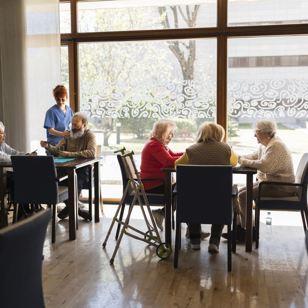 A caregiver assists elderly residents playing cards at tables in a bright, sunlit community room of an assisted living facility.