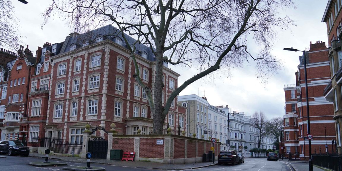  A quiet residential street corner featuring multi-story brick buildings with white-framed windows and ornate architectural details. A large tree and parked cars line the street under an overcast sky, creating a calm urban neighborhood scene.