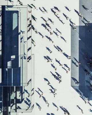 A high-angle view of a bustling urban plaza surrounded by tall, reflective modern buildings. Numerous pedestrians cast long shadows across the open space, creating dynamic patterns in the daylight.