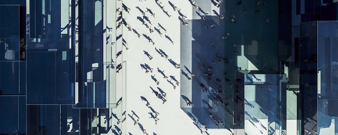 A high-angle view of a bustling urban plaza surrounded by tall, reflective modern buildings. Numerous pedestrians cast long shadows across the open space, creating dynamic patterns in the daylight.