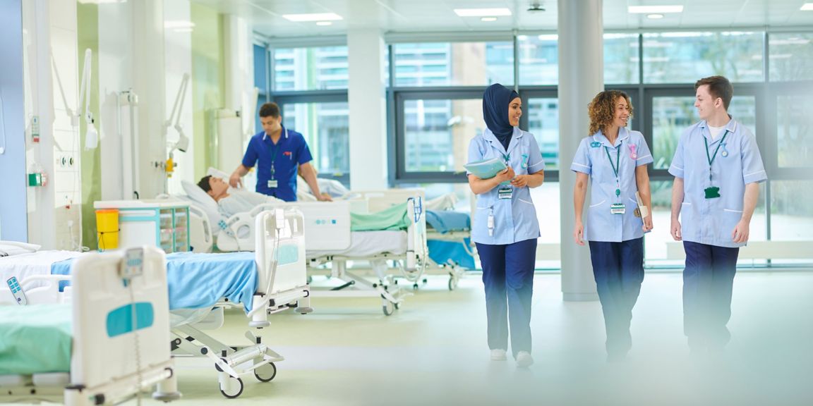 A bright hospital ward featuring healthcare professionals in light blue uniforms attending to patients. The scene includes a patient in bed receiving care, with large windows allowing natural light to illuminate the clinical environment.