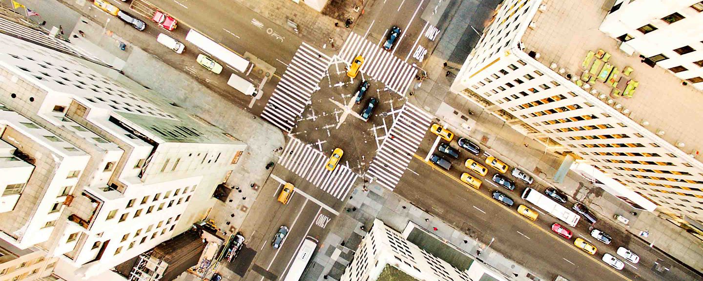 An overhead view of a bustling urban intersection featuring yellow taxis, cars, and buses navigating multiple lanes and crosswalks. Tall buildings surround the area, with rooftops and street markings clearly visible.
