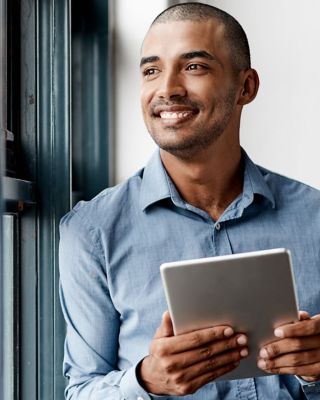 A person in a blue shirt stands by a window, holding and viewing a tablet. The face is blurred for privacy, and the setting suggests a modern, tech-savvy work environment with natural light.