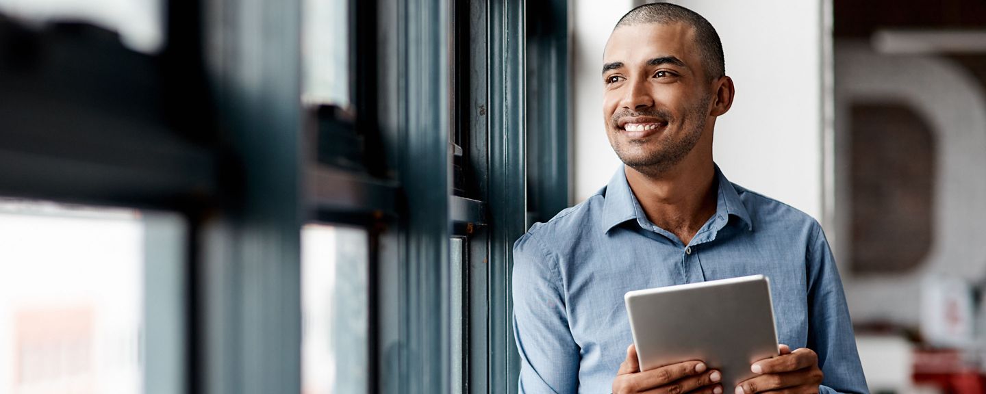 A person in a blue shirt stands by a window, holding and viewing a tablet. The face is blurred for privacy, and the setting suggests a modern, tech-savvy work environment with natural light.