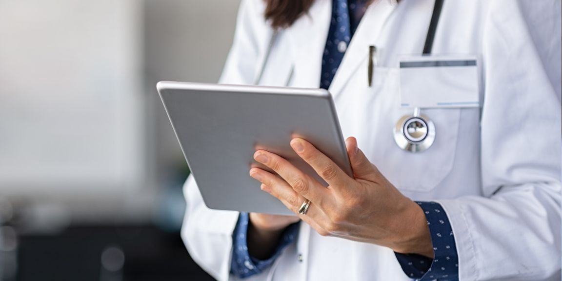Close-up of a doctor in a white coat holding a tablet, with a stethoscope and ID badge visible.