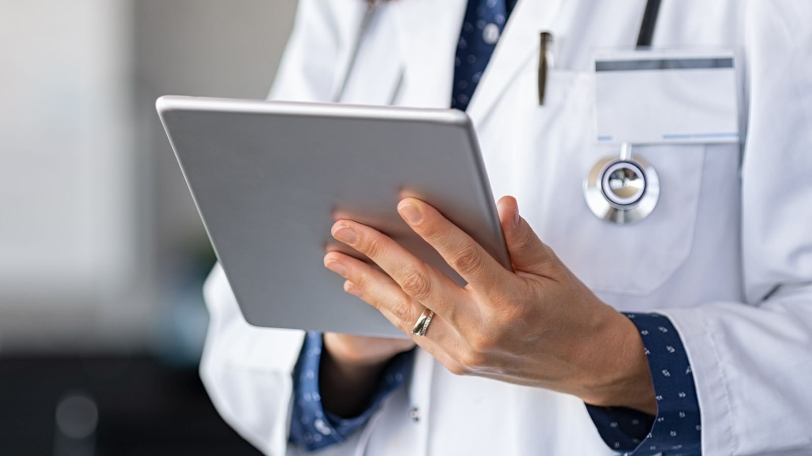 Close-up of a doctor in a white coat holding a tablet, with a stethoscope and ID badge visible.