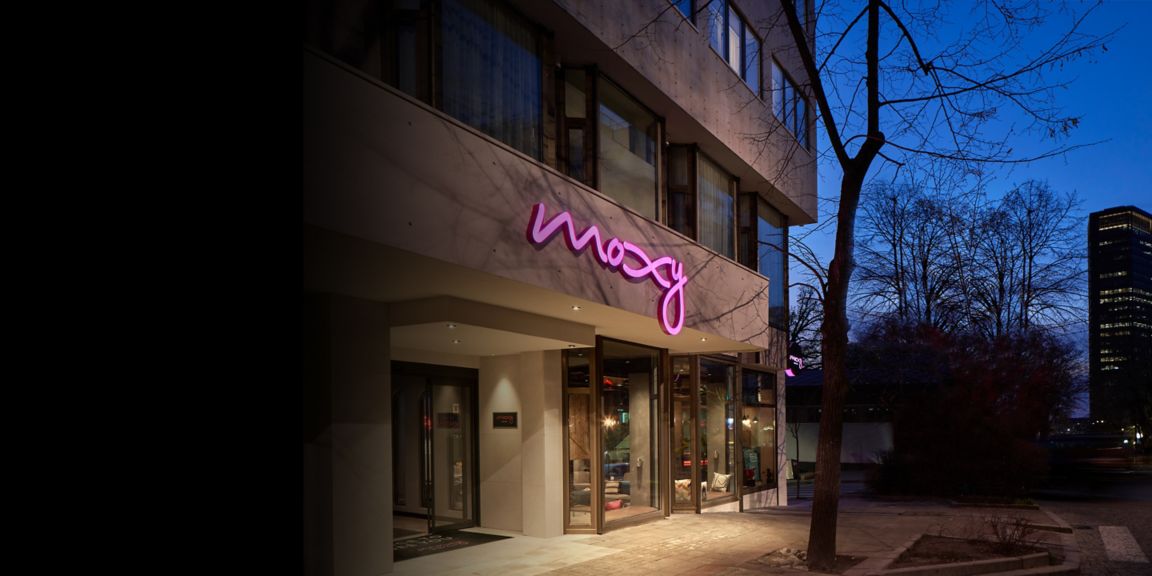 Street‑level façade featuring illuminated lettering above glass doors, sidewalk lighting, and surrounding urban buildings during early evening.