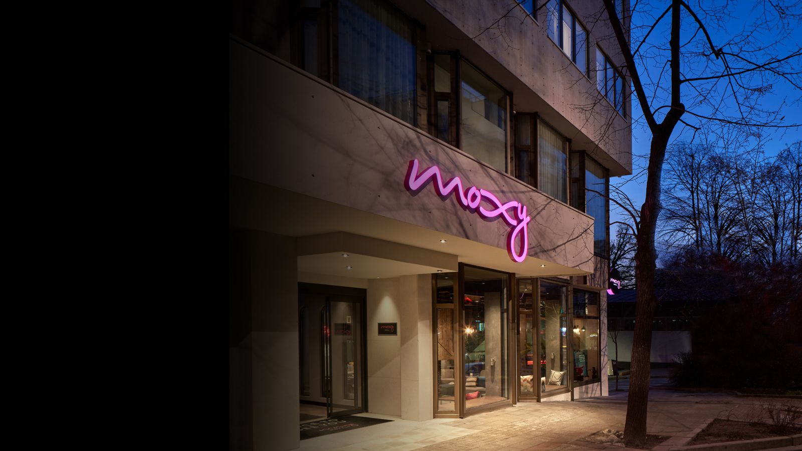 Street‑level facade featuring illuminated lettering above glass doors, sidewalk lighting, and surrounding urban buildings during early evening.