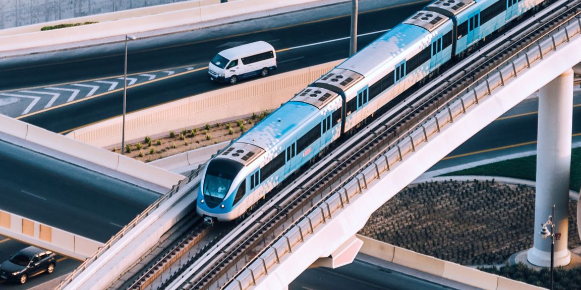 A sleek blue and white elevated train crosses a concrete bridge above a busy road with a white van below. Ideal for showcasing urban transportation, infrastructure, or smart city mobility solutions.