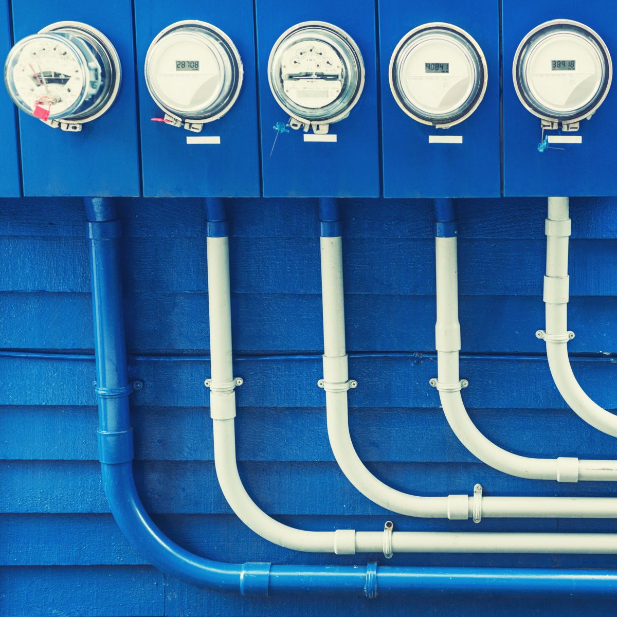 A row of electric meters mounted on a vibrant blue wall with neatly routed conduits.
