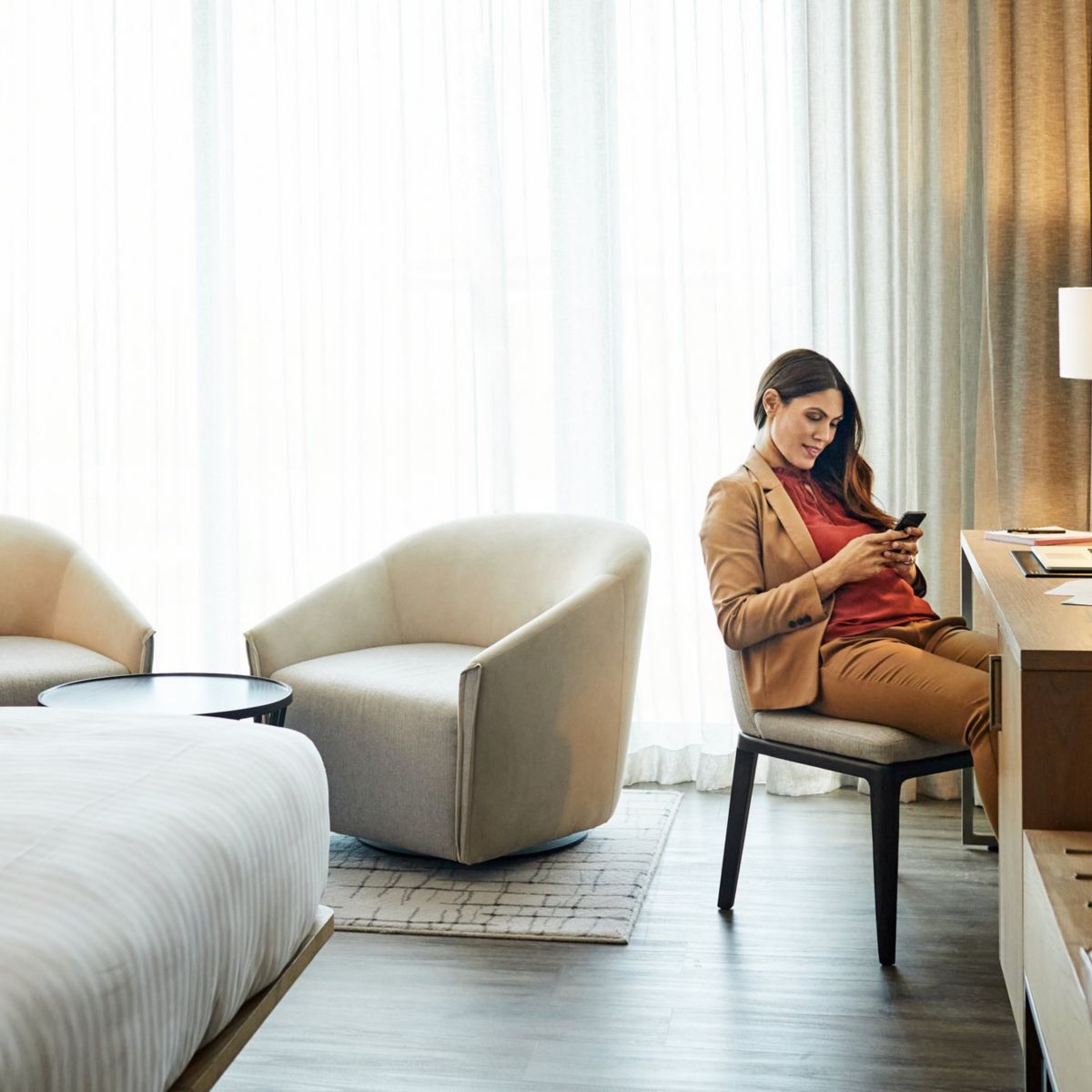 A professional woman sits at a hotel room desk using her smartphone while working on her laptop during a business trip.