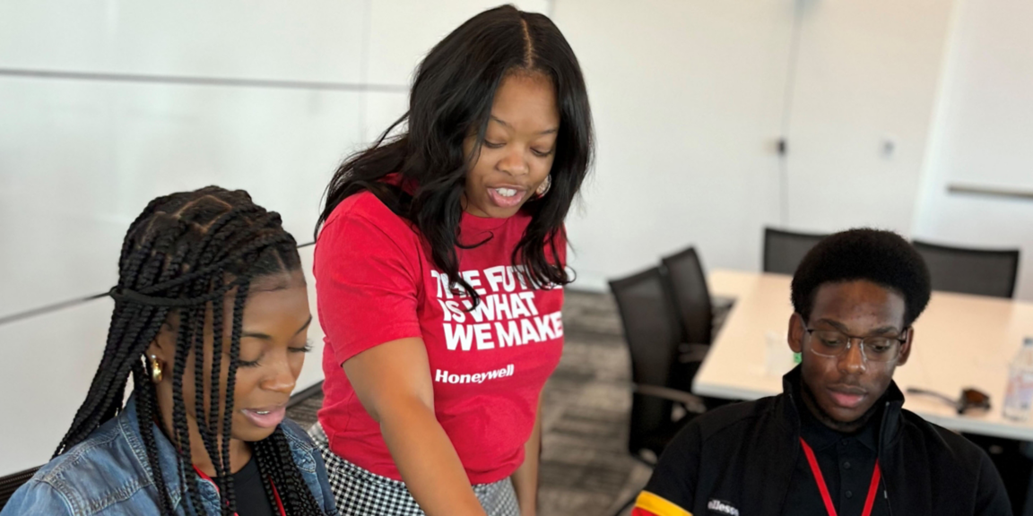 Mentor standing beside two students during a hands-on learning session in a meeting room, reviewing work together at a table.