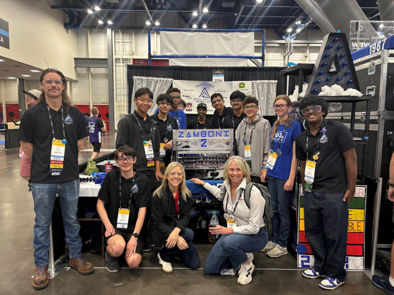 Group of students and mentors posing with a robotics machine at a robotics competition booth inside a large convention center.