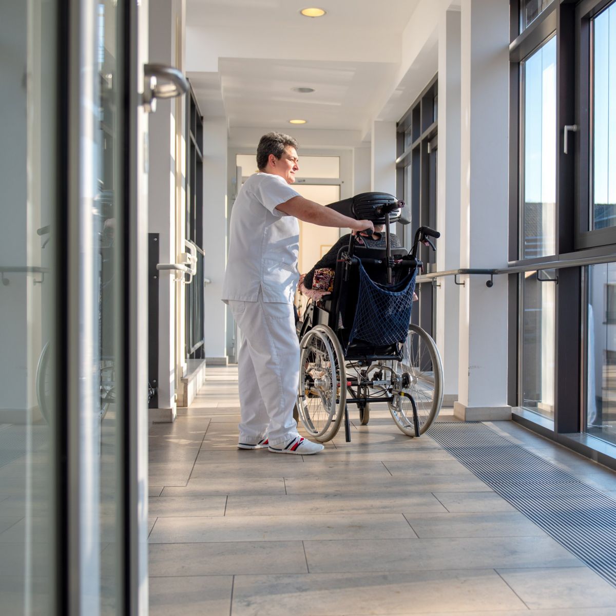 A caregiver in white scrubs pushes a patient in a wheelchair down a bright, sunlit corridor in a modern medical or nursing facility.