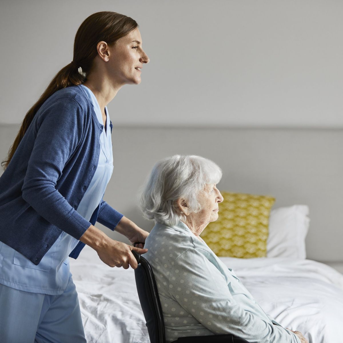 A professional caregiver assists an elderly woman in a wheelchair inside a comfortable, modern bedroom setting.