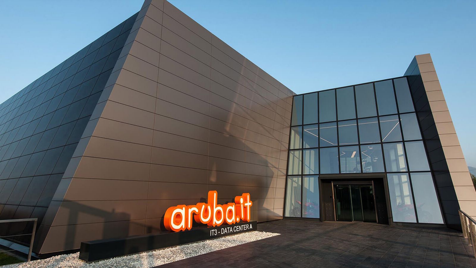 Exterior of modern building with glass facade entrance, paved walkway, and illuminated aruba.it sign on the wall.