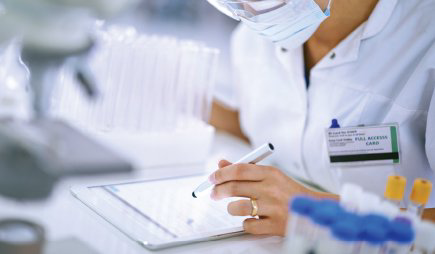 A healthcare professional writes on a clipboard beside laboratory samples and an identification badge.