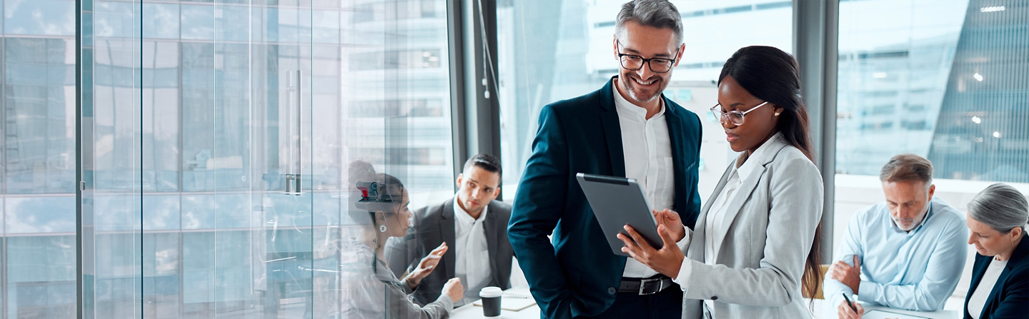 Two business colleagues review a tablet while other professionals meet in a glass-walled office.
