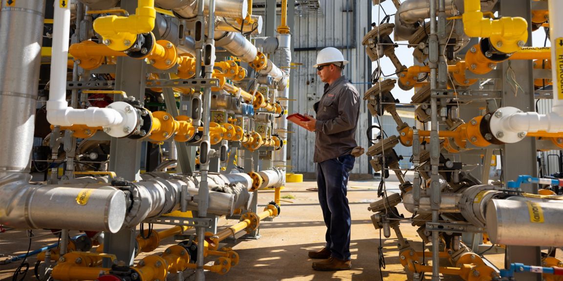 A technician wearing a hard hat stands in a complex industrial environment, holding a red tablet while inspecting machinery, pipes, and valves. The image conveys themes of safety, maintenance, and operational oversight.