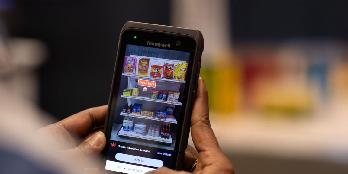 A person uses a Honeywell device to check store inventory, displaying an "Out Of Stock" alert on snacks and beverages.