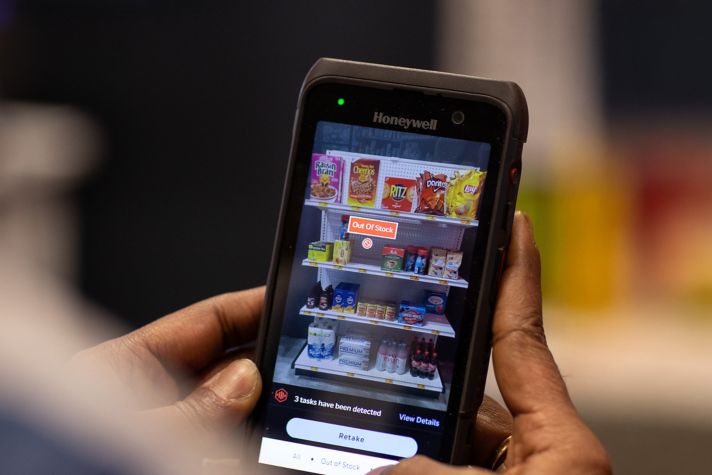 A person uses a Honeywell device to check store inventory, displaying an "Out Of Stock" alert on snacks and beverages.