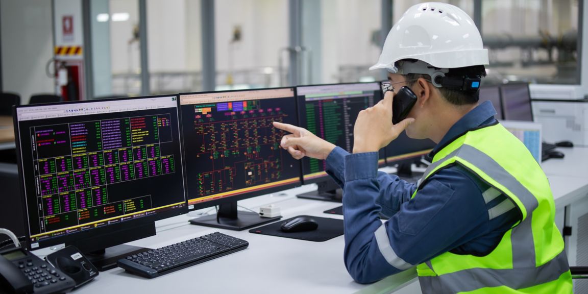 A technician in safety helmet uses computer systems to control and monitor substation operations with multiple screens displaying data and control panels in industrial setting