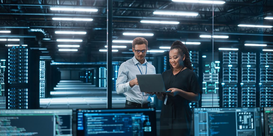 Two professionals standing in a modern data center environment review information on a laptop, surrounded by rows of illuminated server racks and multiple screens displaying system data and code interfaces.
