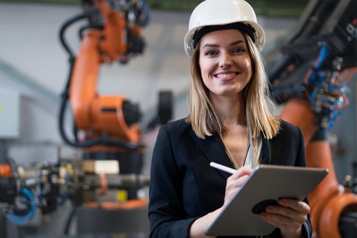 A professional woman wearing a hard hat is working with robotic systems in an industrial environment, showcasing the integration of technology in modern manufacturing.